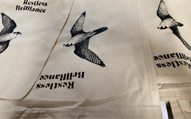 Canvas tote bags with a peregrine falcon and 'Restless Brilliance' printed on it.