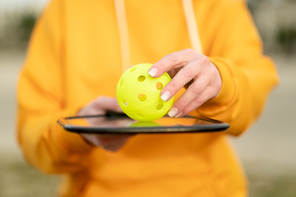 Close up of woman in an orange jumper holding flat racket with a yellow ball with holes in, on top 