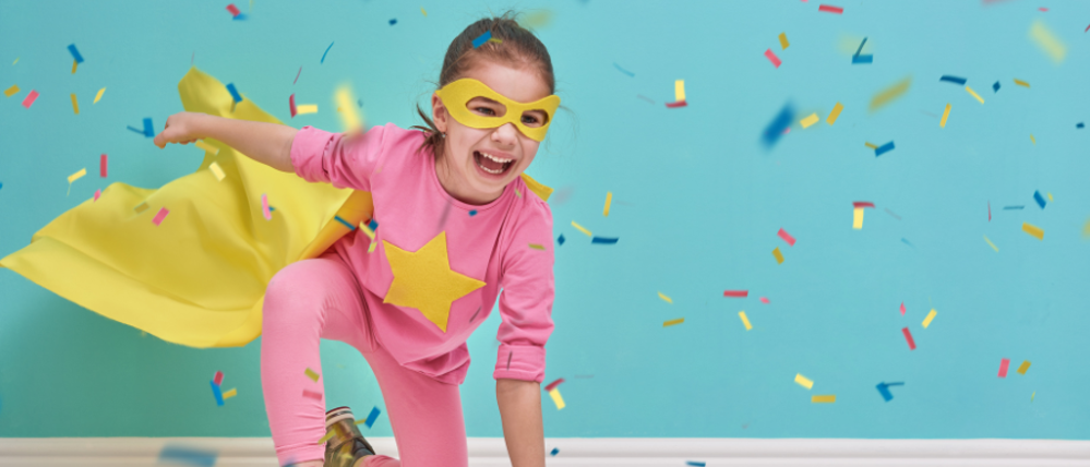 Girl dressed as superhero, wearing pink outfit and yellow cape and mask, with confetti tumbling around her