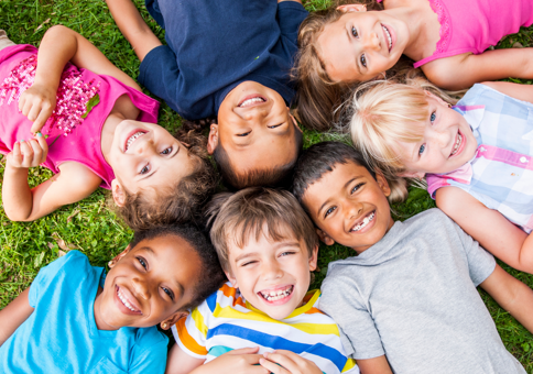 Group of happy young children lying on the grass in the sunshine a circle with their heads in the middle.