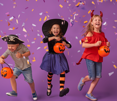 Kids in Halloween fancy dress jumping and smiling in front of confetti and a purple background.