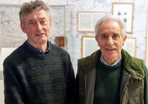 Two men in jumpers smiling at the camera. Behind them are maps on the wall and diaries of J.A. Baker on display. 