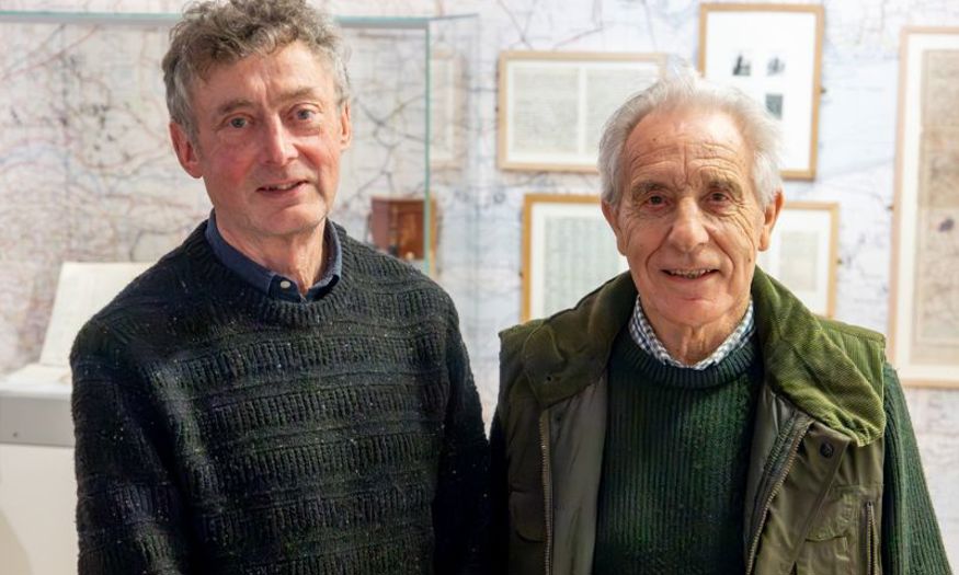 Two men in jumpers smiling at the camera. Behind them are maps on the wall and diaries of J.A. Baker on display. 