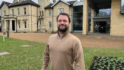 A man standing on the lawn outside Chelmsford Museum smiles at the camera. 