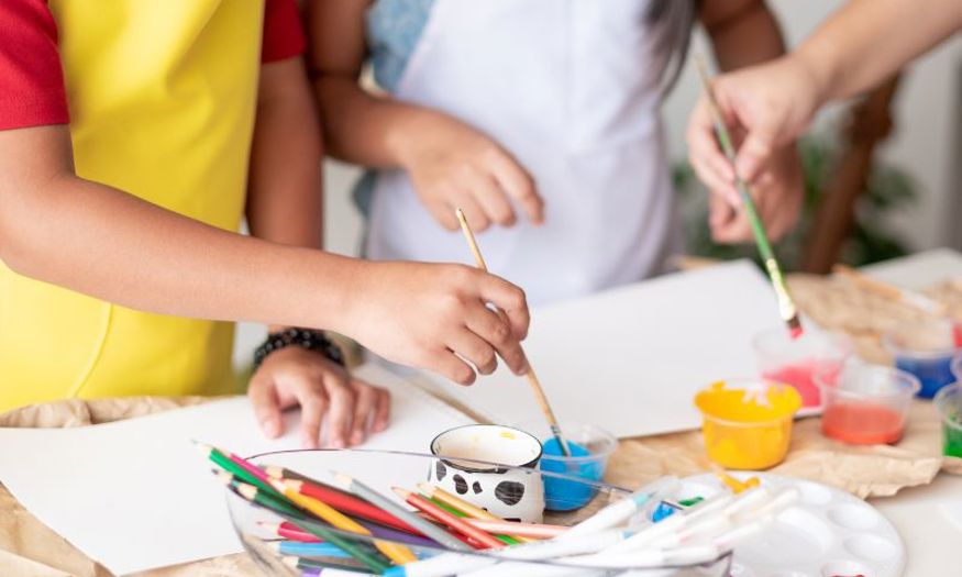 Children using paint to create art on white paper. Beside them is a container filled with coloured pencils and pens. 