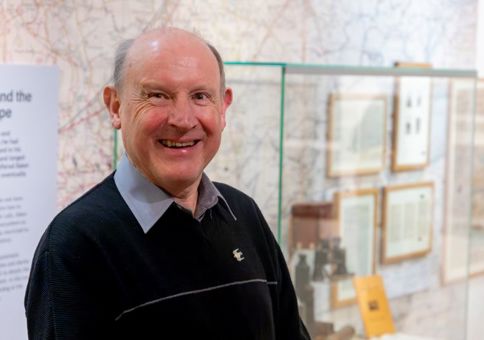 David Simmonds smiling inside the J.A. Baker exhibition. Behind him is a glass case filled with Baker's items, such as binoculars and journals. 