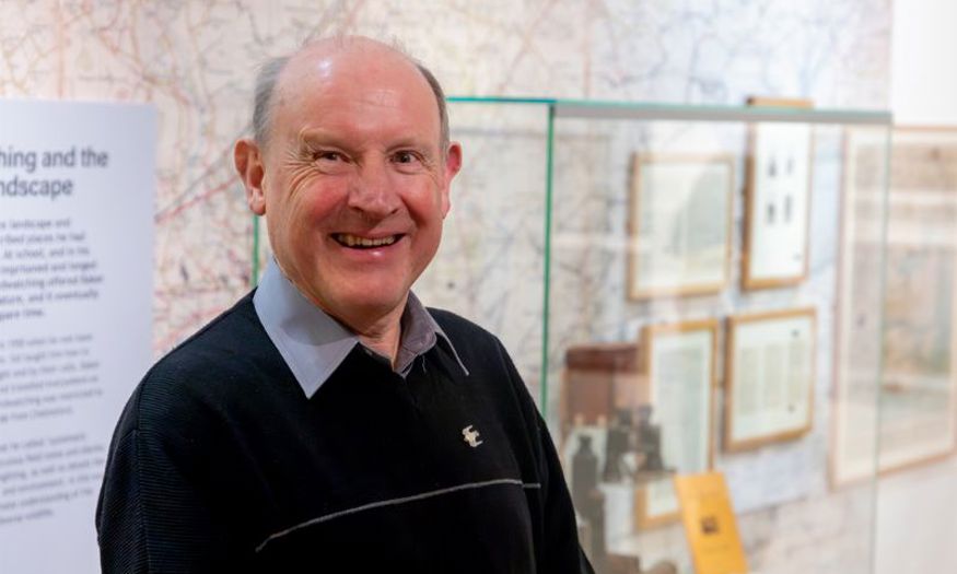 David Simmonds smiling inside the J.A. Baker exhibition. Behind him is a glass case filled with Baker's items, such as binoculars and journals. 