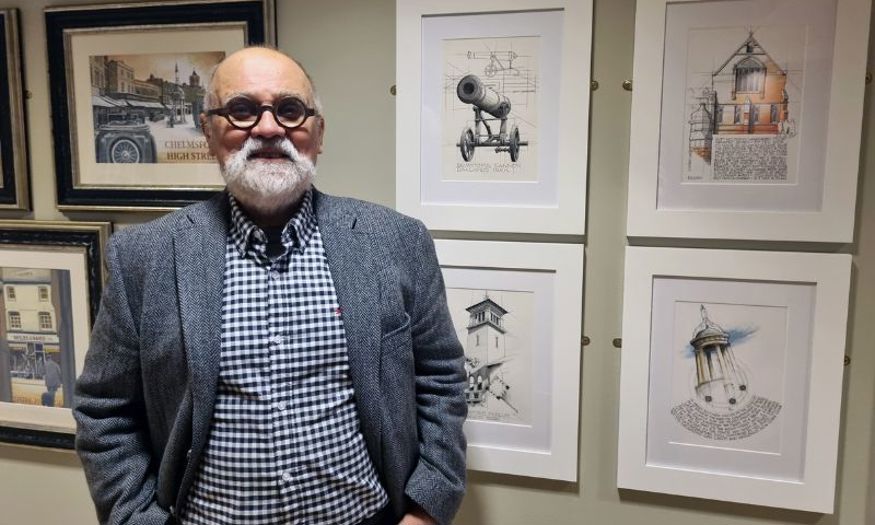 An older man with grey hair and dark circular glasses stands in front of a wall with various framed artwork hung. 