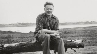 A black and white photo of JA Baker sitting on a log by the waterside. 