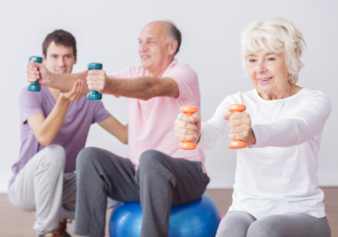 Man coaching older people who are sitting on exercise balls and using hand weights