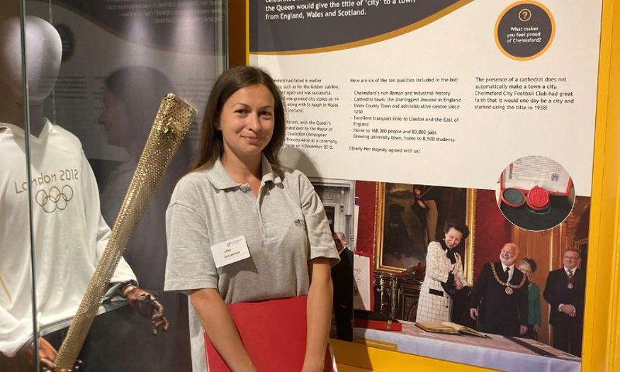 A woman with long, dark hair smiling in front of a London Olympics 2012 uniform on display. 