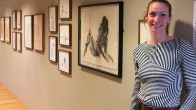 A woman with a brunette ponytail and striped shirt stands in front of a wall covered in framed black and white artwork. 