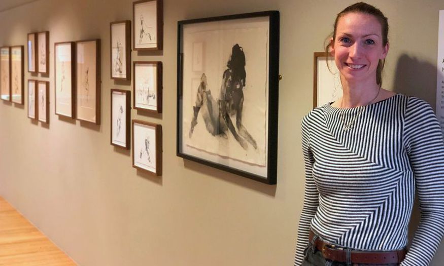 A woman with a brunette ponytail and striped shirt stands in front of a wall covered in framed black and white artwork. 