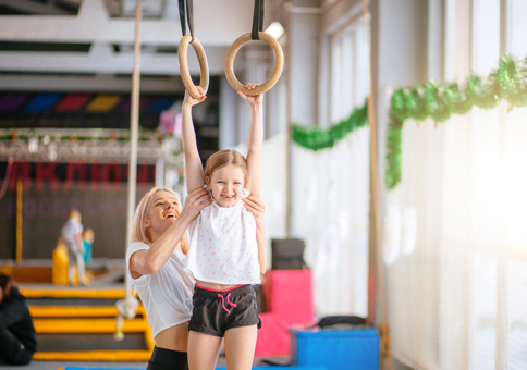 Young girl smiling being held up on gymnastics rings by a gymnastics teacher 