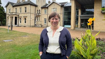 A woman with short brown hair smiles at the camera while stood outside of Chelmsford Museum. 