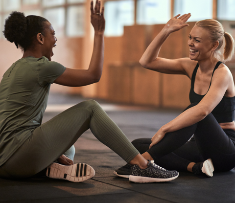 Two women it fitness clothes sitting on floor of exercise studio, about to give each other a high-five