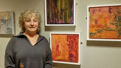 A woman with chin-length hair smiles in front of a wall with colourful pieces of art hanging on it. 