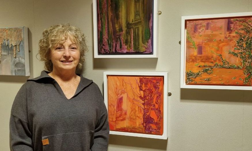 A woman with chin-length hair smiles in front of a wall with colourful pieces of art hanging on it. 